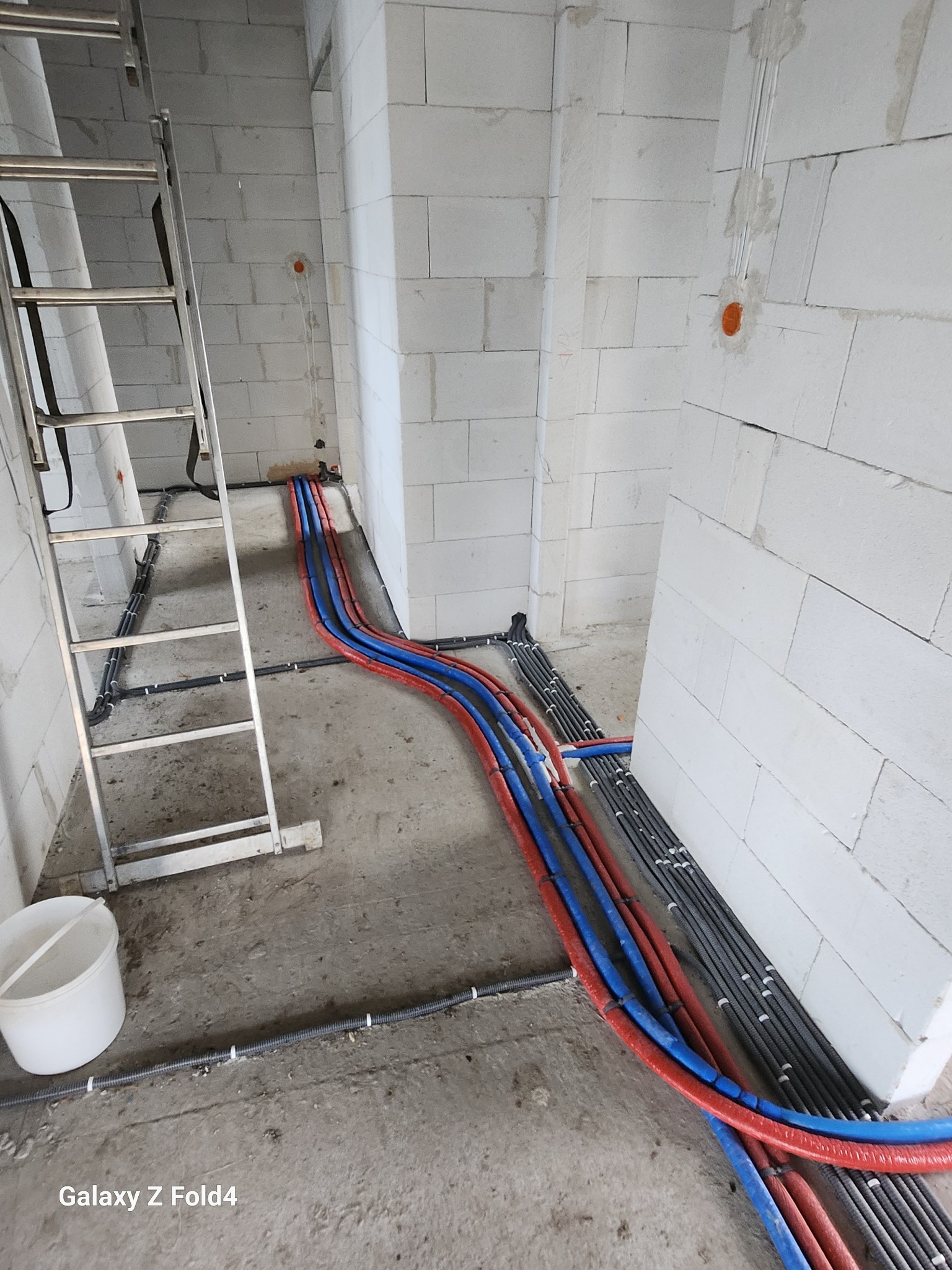 Instalacja rur grzewczych (czerwone i niebieskie) i przewodów elektrycznych w peszlach na surowym podłożu w nowym budynku z białych bloczków. Widoczna drabina i wiadro.