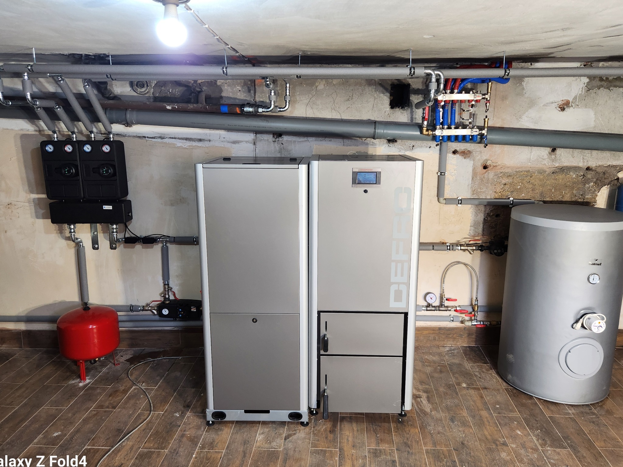 Kocioł grzewczy z podajnikiem i zasobnikiem wody użytkowej, zainstalowany w pomieszczeniu z widoczną instalacją hydrauliczną i elementami sterującymi.