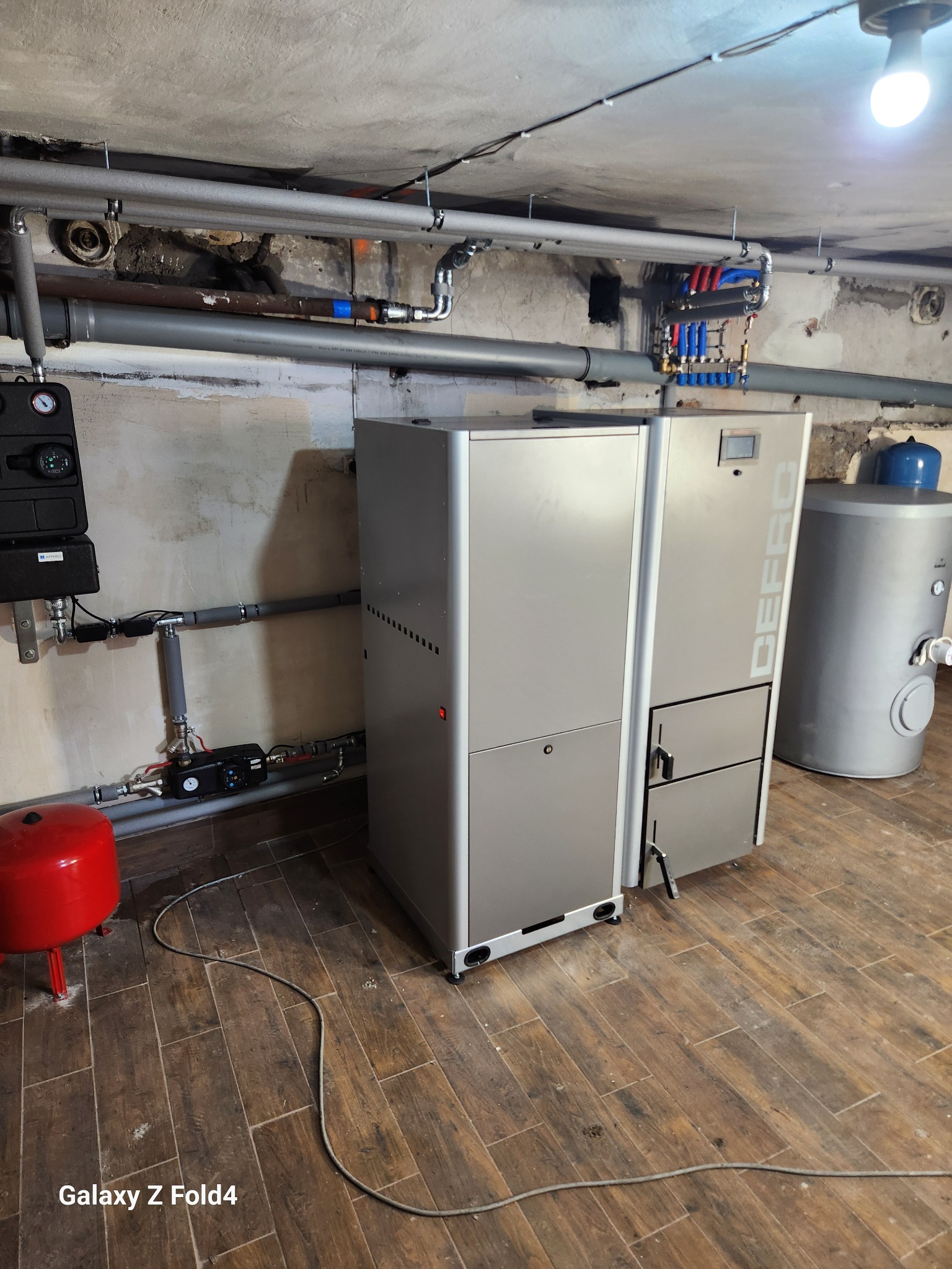 Kotłownia z nowoczesnym piecem Defro, zasobnikiem ciepłej wody i instalacją grzewczą. Rury miedziane i stalowe, manometry, brązowe płytki na podłodze.