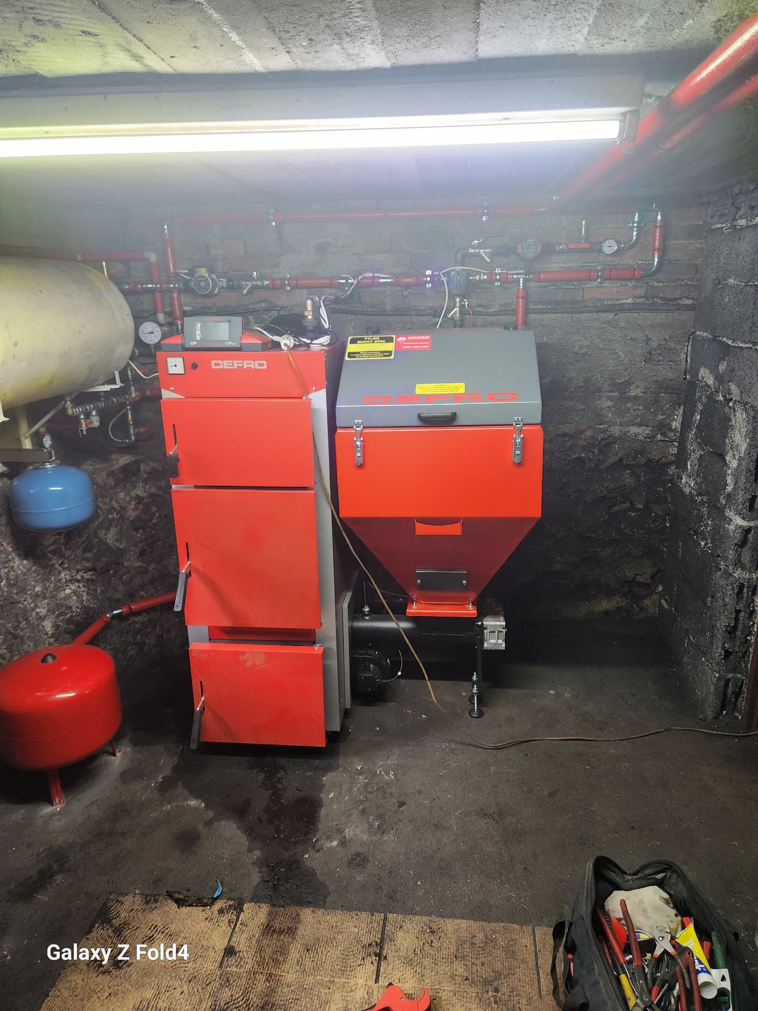 Czerwony piec CO marki Defro w kotłowni, z widocznymi rurami instalacji grzewczej i torbą narzędziową w rogu. Surowe, ciemne otoczenie.