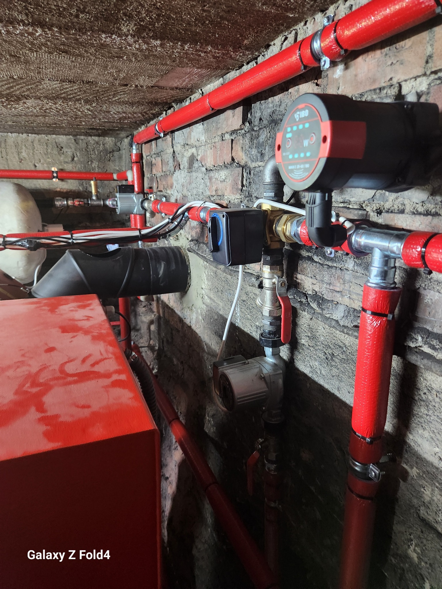 Czerwone rury instalacji CO z pompą obiegową i zaworami, zamontowane w piwnicy z ceglanym murem. Instalacja połączona z kotłem grzewczym.