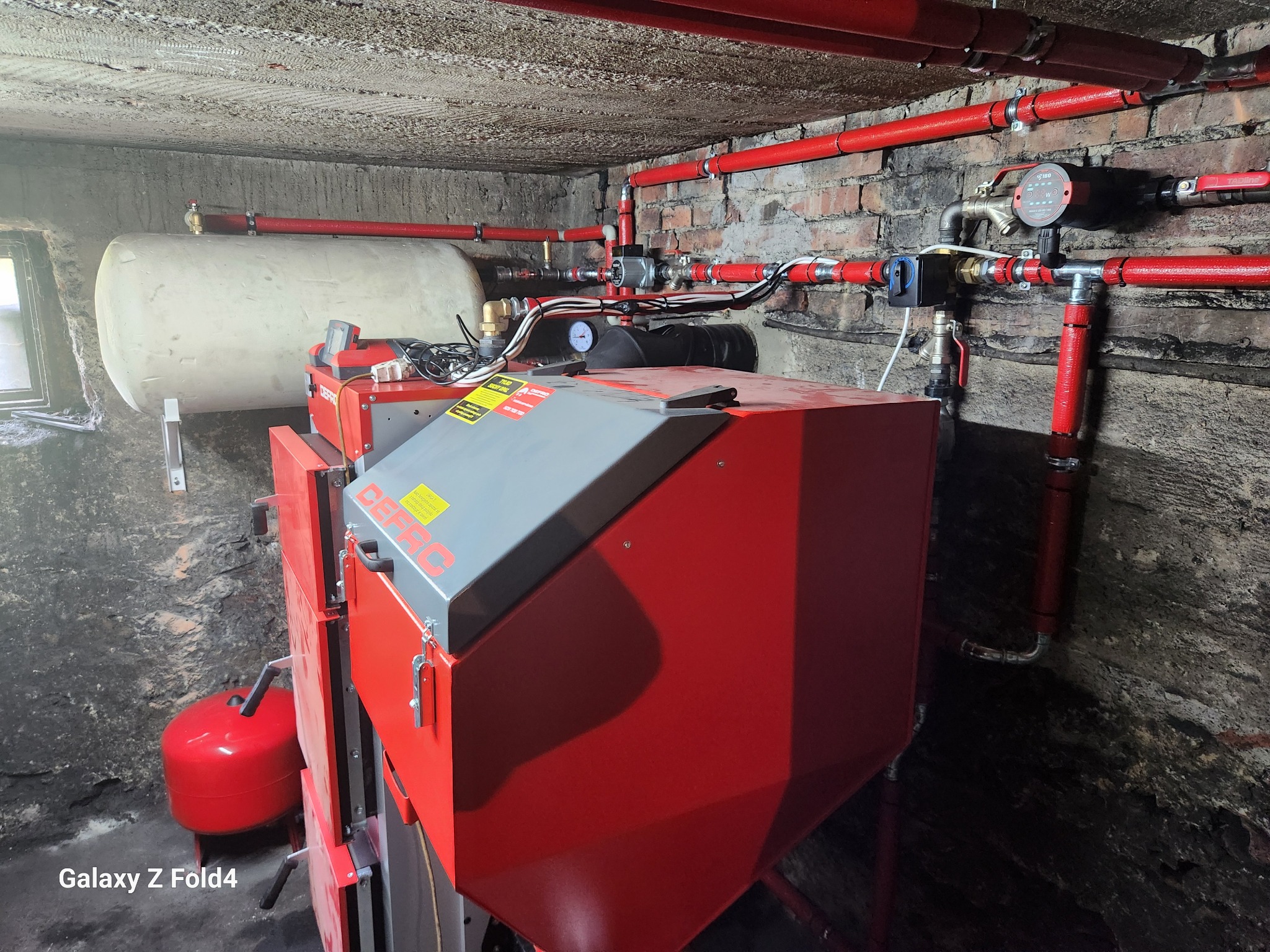 Czerwony piec CO marki DEFRO z szarym blatem, zbiornik wyrównawczy i instalacja grzewcza z rurami w piwnicy o surowych ścianach z cegły i betonu.