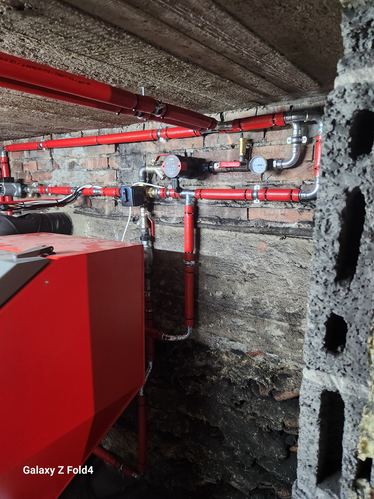 Czerwone rury instalacji grzewczej zamontowane na ceglanej ścianie w piwnicy, z widocznym kotłem i manometrami. Instalacja wykonana w surowym wnętrzu.