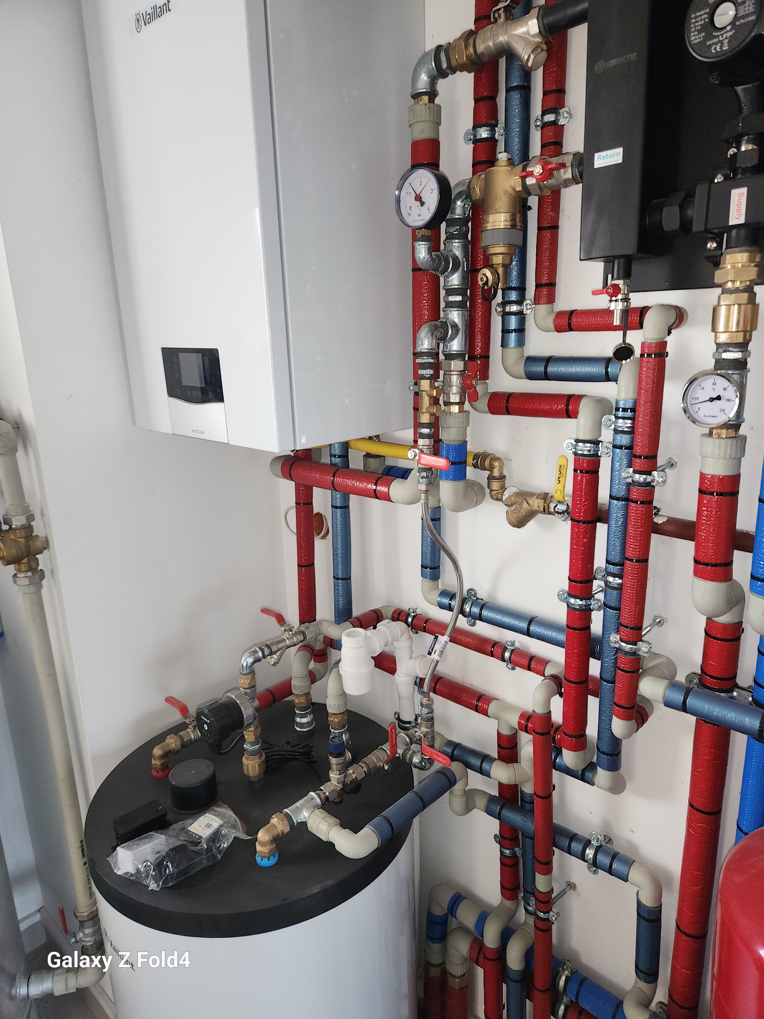 Biały piec Vaillant i rozbudowana instalacja hydrauliczna z rurami w izolacji czerwonej i niebieskiej, manometry, zawory. Widok z góry na zbiornik.