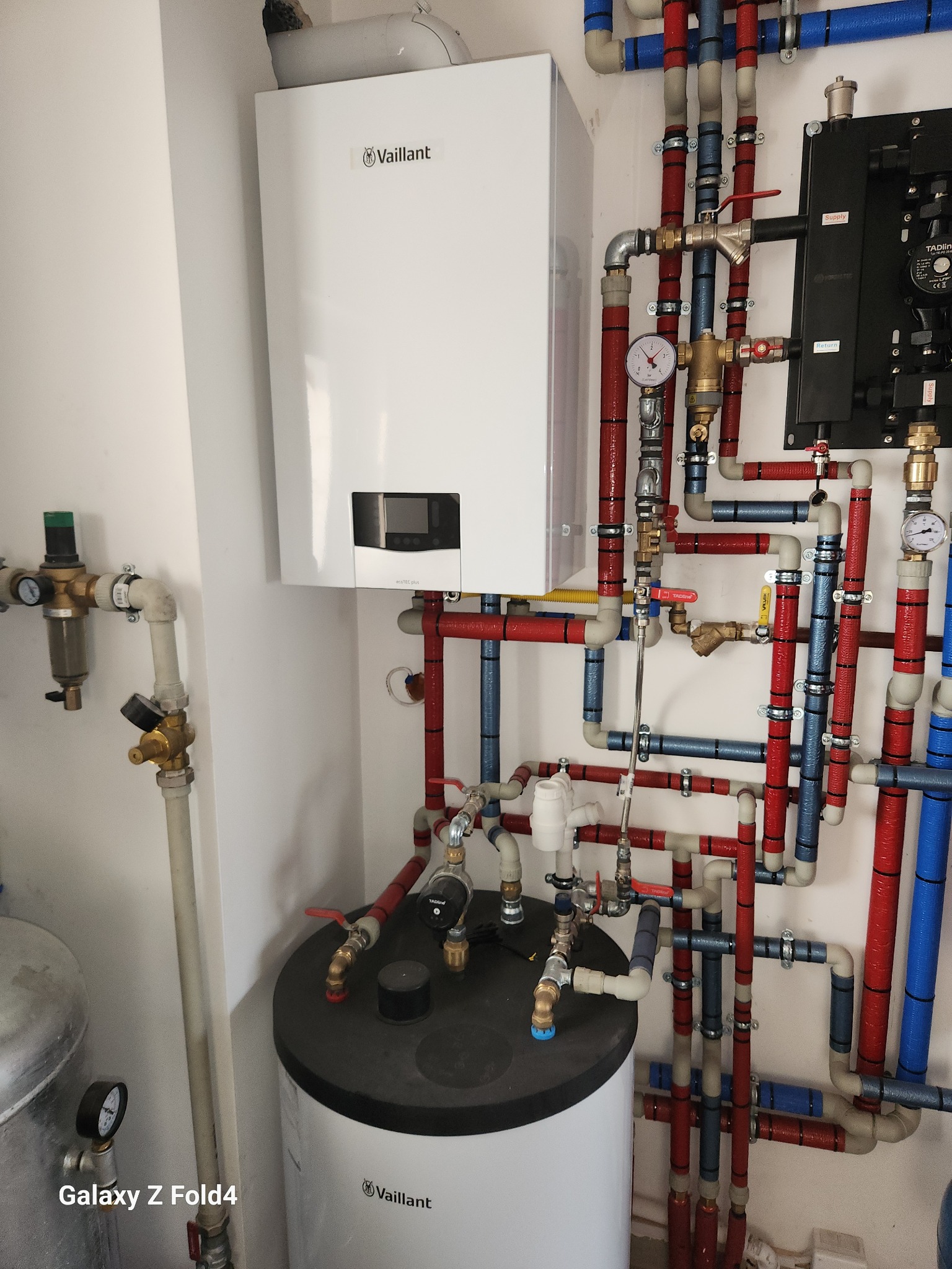 Biały piec Vaillant z rozbudowaną instalacją hydrauliczną w kolorze czerwonym i niebieskim. Widoczne zawory, manometry i zbiornik.