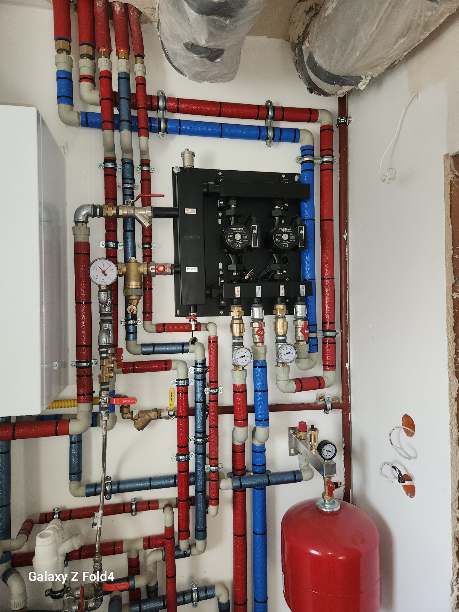 Kompleksowa instalacja grzewcza z rurami w kolorach czerwonym i niebieskim, zaworami, manometrami i czarnym rozdzielaczem na białej ścianie w trakcie montażu.
