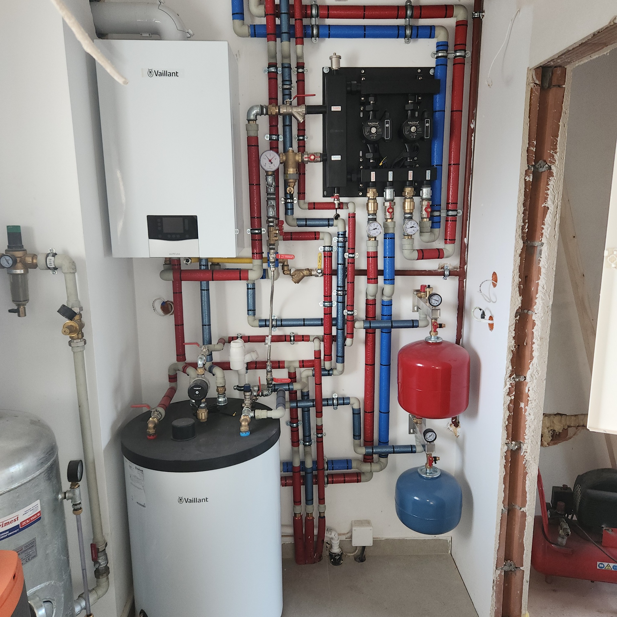 Kompletna instalacja grzewcza z kotłem Vaillant, zbiornikiem, rozdzielaczem i rurami w kolorach czerwonym i niebieskim. Widoczne manometry i zawory.