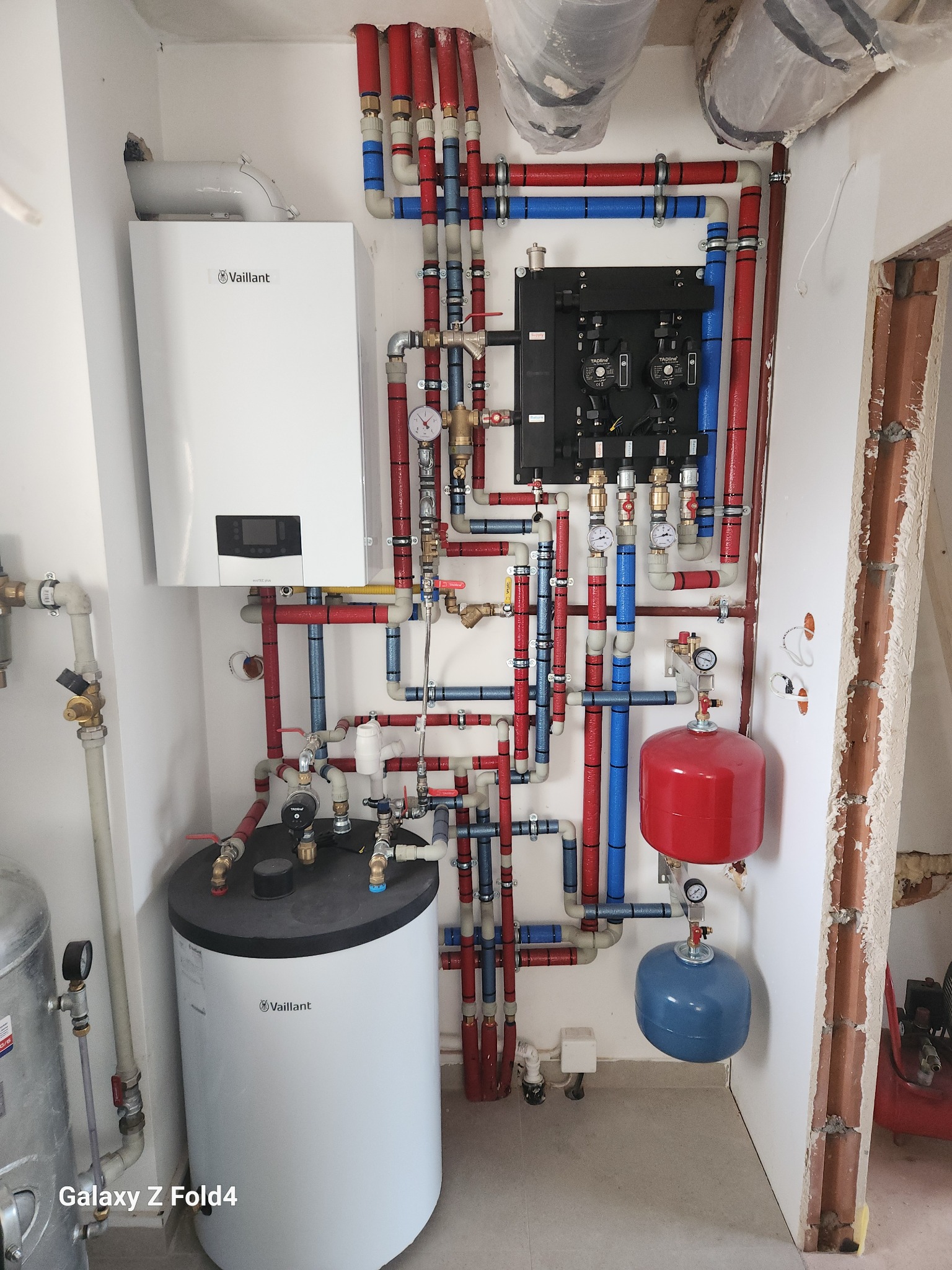 Złożona instalacja grzewcza z kotłem Vaillant, zbiornikiem i rurami w kolorach czerwonym i niebieskim, zamontowana na białej ścianie w kotłowni.