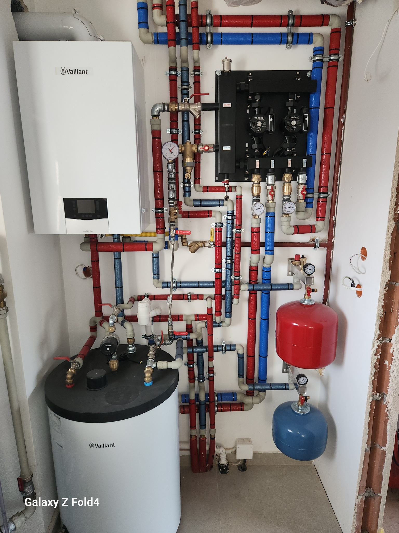 Kompleksowa instalacja grzewcza Vaillant z czerwono-niebieskimi rurami, kotłem, zasobnikiem i czarną rozdzielnicą, zamontowana na białej ścianie w surowym stanie.