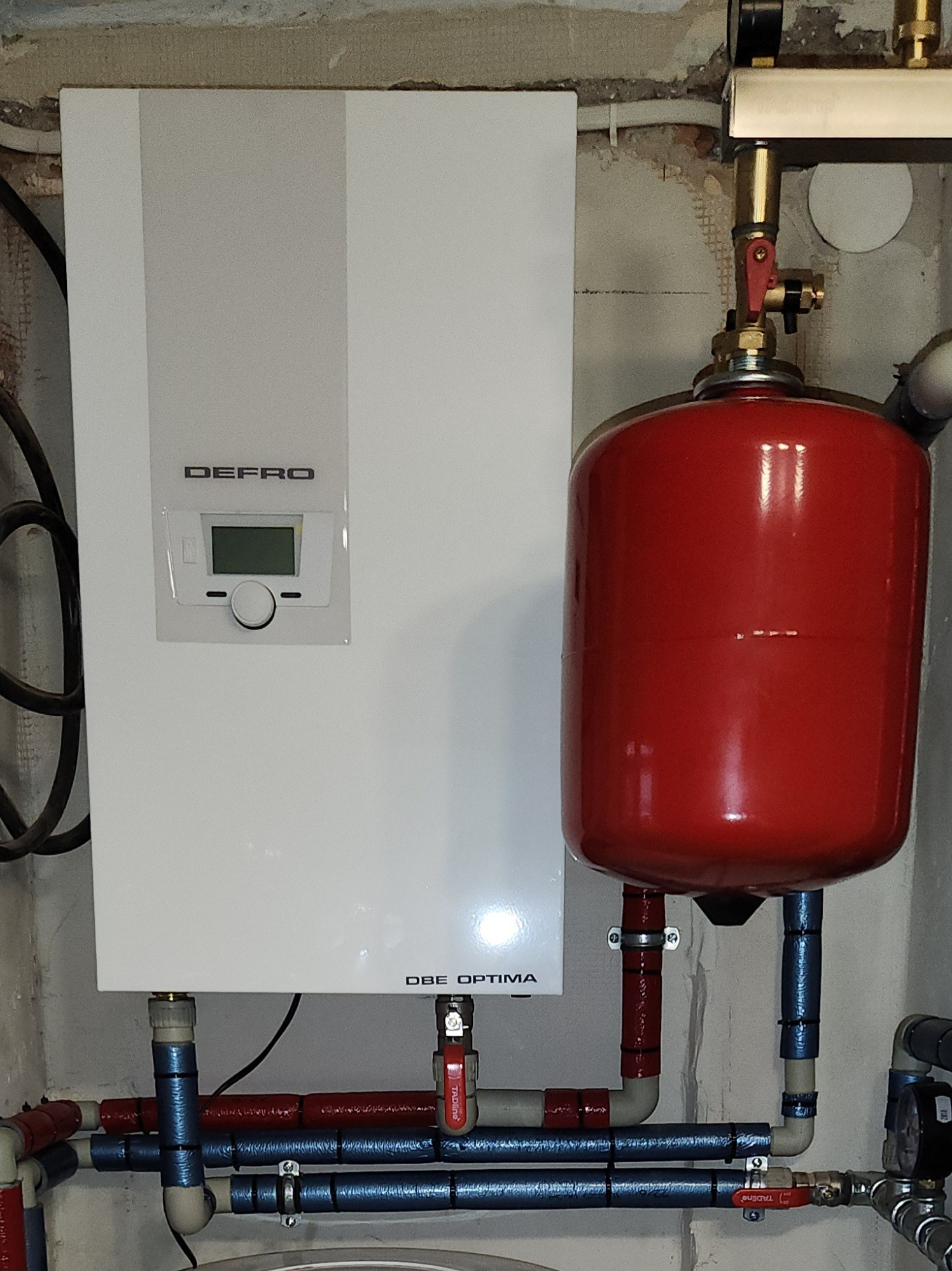 Biały piec grzewczy Defro Optima z czerwonym zbiornikiem wyrównawczym i rurami w izolacji, zamontowany na tle surowej ściany. Widoczne zawory i przyłącza.