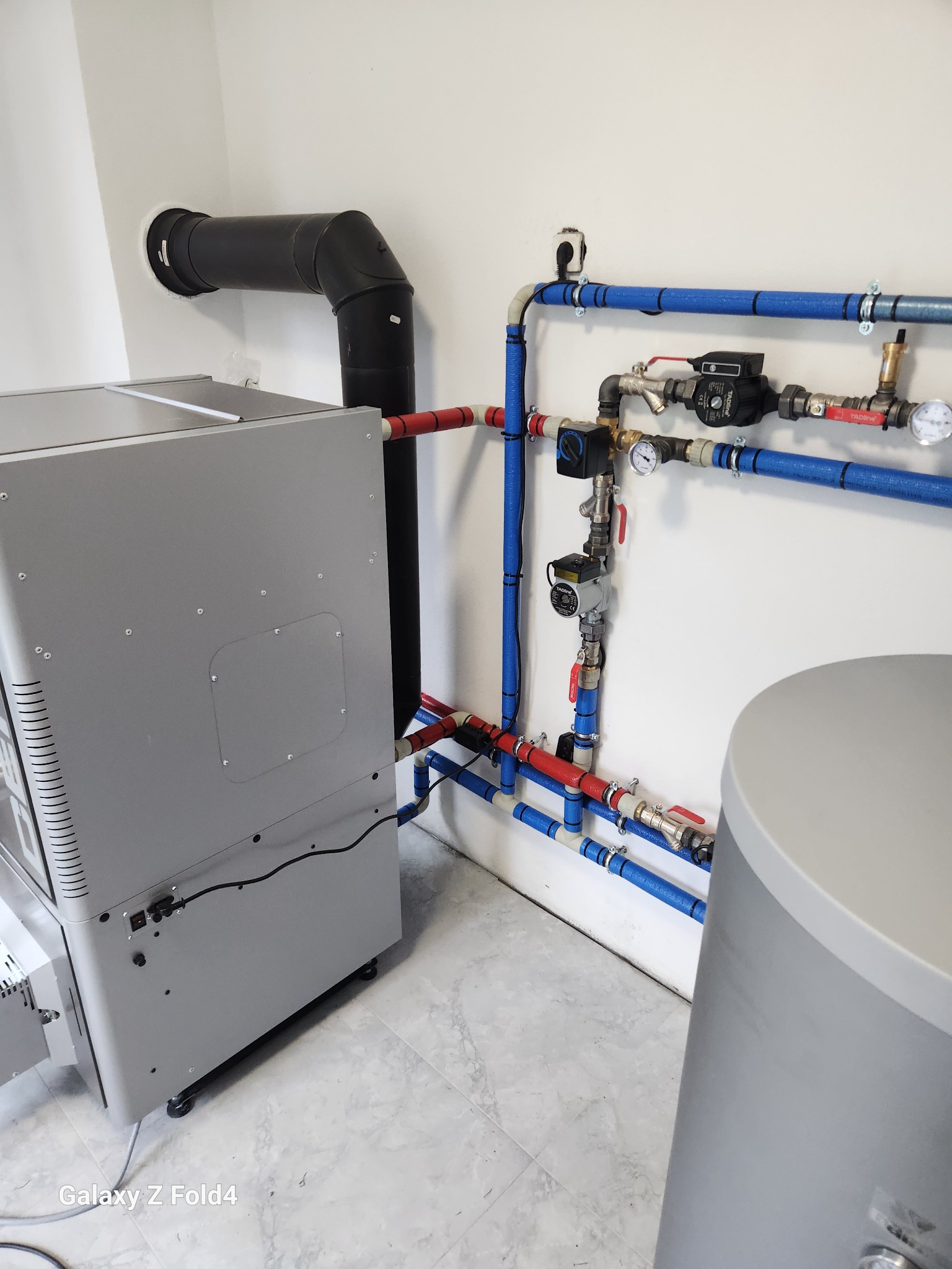 Nowoczesna kotłownia: piec z czarnym kominem, niebieskie i czerwone rury instalacji grzewczej, zbiornik. Instalacja po montażu, gotowa do użytku.