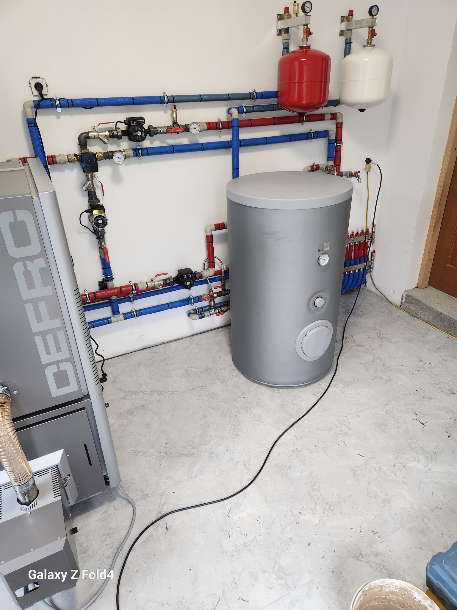 Kotłownia z kotłem Defro i zbiornikiem Galmet, instalacja z czerwono-niebieskimi rurami, manometry, zawory, zbiorniki wyrównawcze. Widok na podłączenia i elementy instalacji.