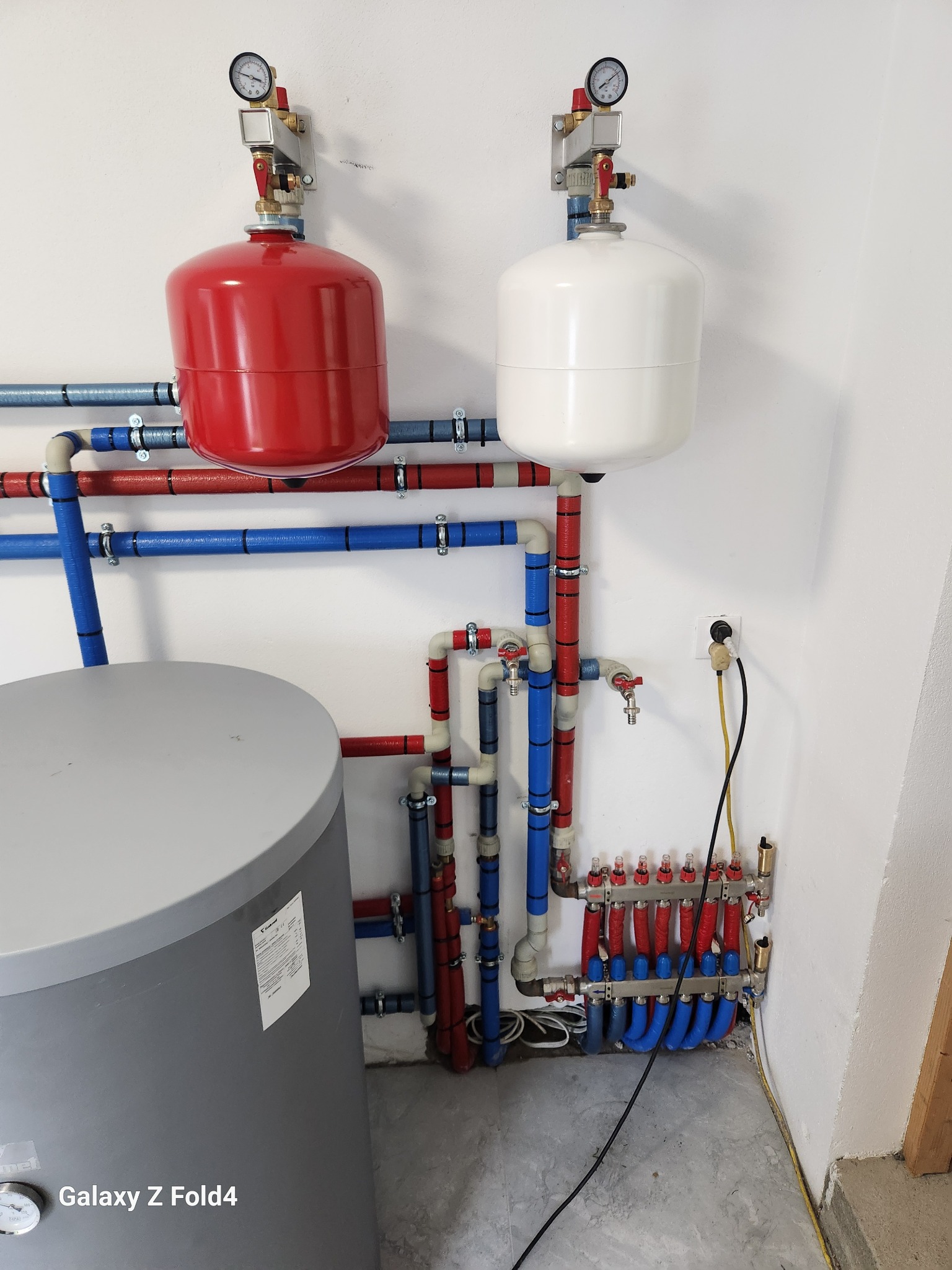 Złożona instalacja grzewcza z czerwonym i białym zbiornikiem wyrównawczym, rurami w kolorach czerwonym i niebieskim oraz szarym zasobnikiem wody w kotłowni.