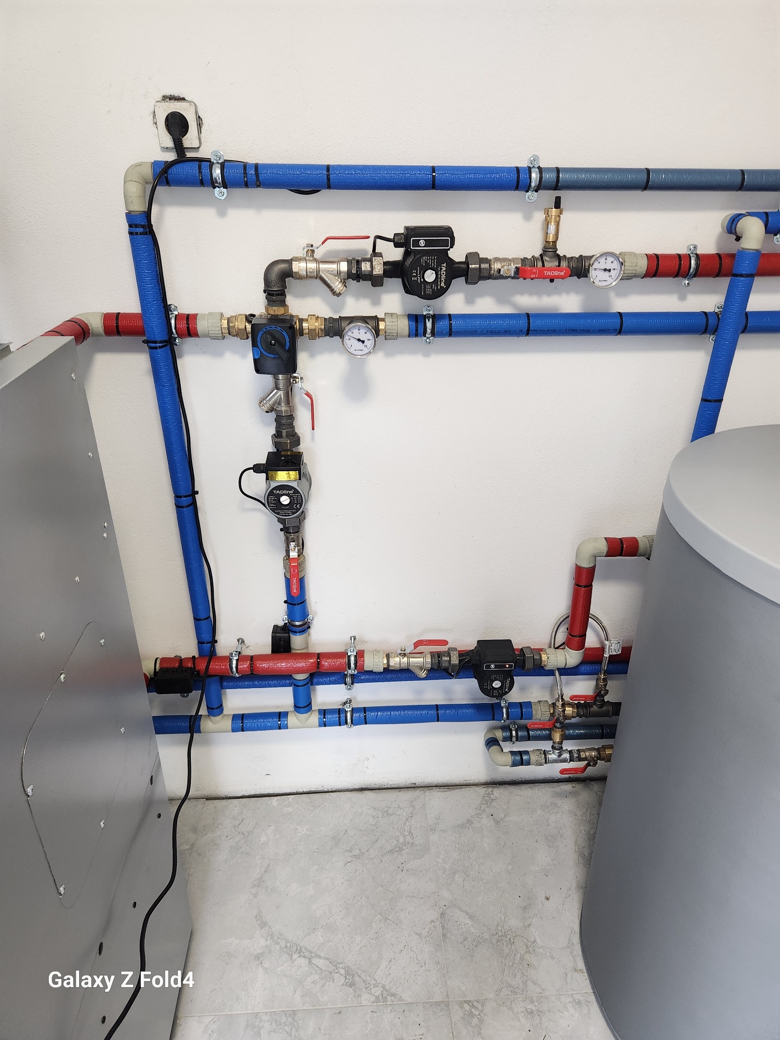 Kompleksowa instalacja hydrauliczna z rurami w izolacji, zaworami, manometrami i pompami obiegowymi w kotłowni. Widoczny zasobnik ciepłej wody i piec.