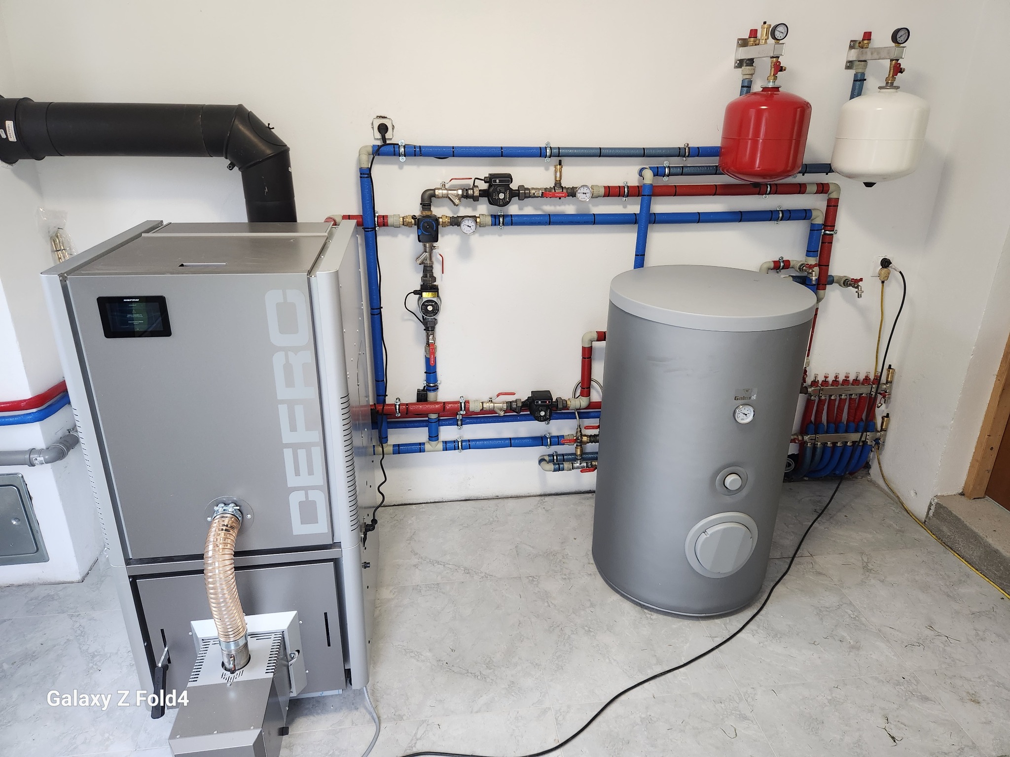 Nowoczesna kotłownia z kotłem na pellet, zasobnikiem ciepłej wody i rozbudowaną instalacją hydrauliczną z czerwono-niebieskimi rurami na białej ścianie.