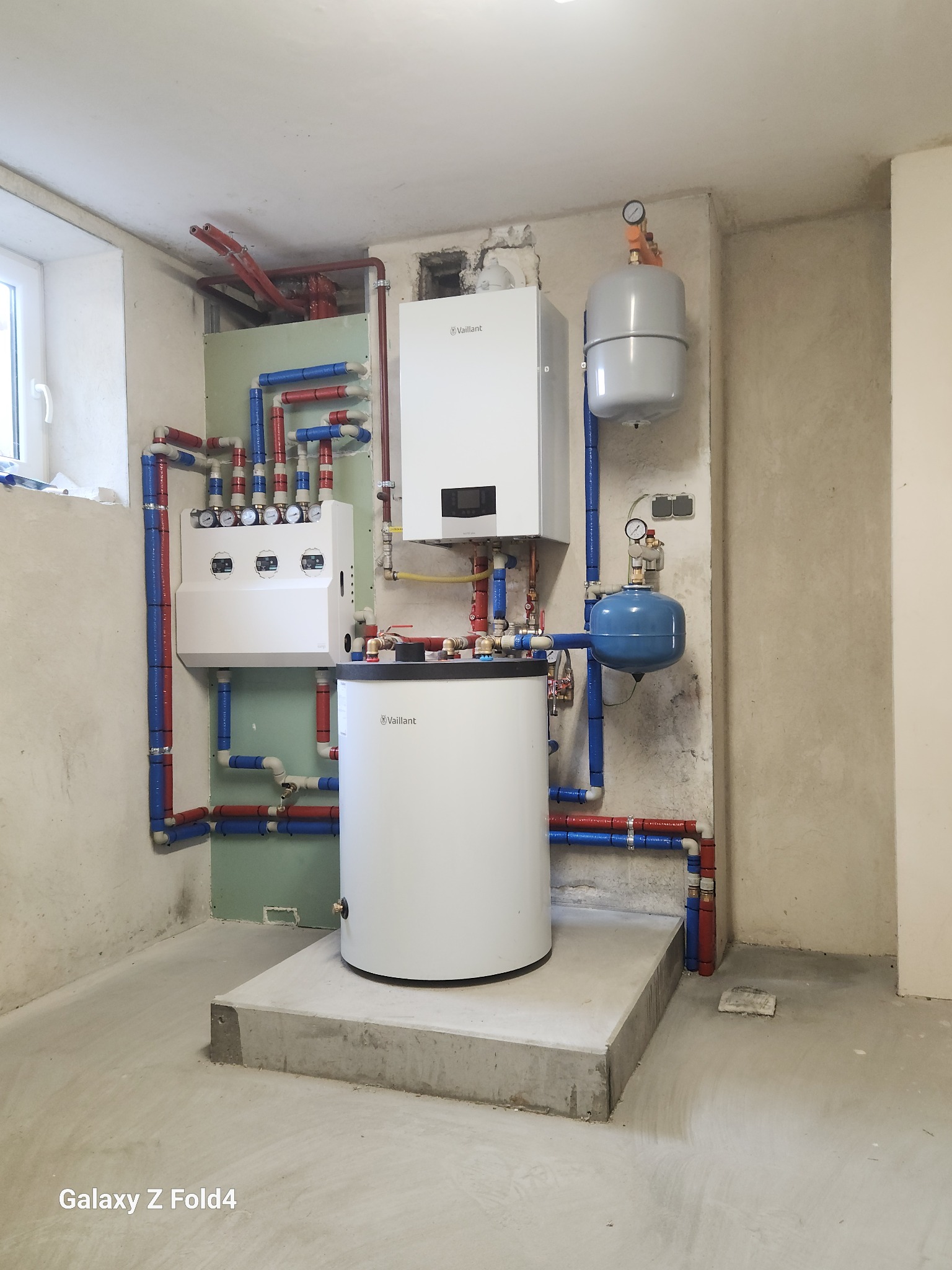 Biały piec Vaillant z zbiornikiem i rozdzielaczem, połączony rurami w kolorach czerwonym i niebieskim. Instalacja w surowym pomieszczeniu z betonową podłogą.