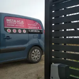 Błękitny samochód dostawczy z reklamą usług elektrycznych i alarmowych firmy Mar-Instal, zaparkowany obok czarnego, nowoczesnego ogrodzenia z zamontowanym napędem bramy.