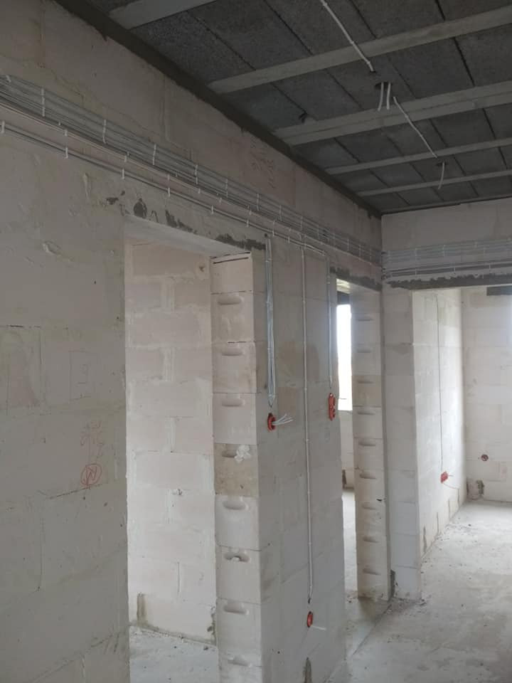 Surowe wnętrze w trakcie budowy z widocznymi przewodami elektrycznymi poprowadzonymi pod sufitem i wzdłuż ścian z betonu komórkowego.
