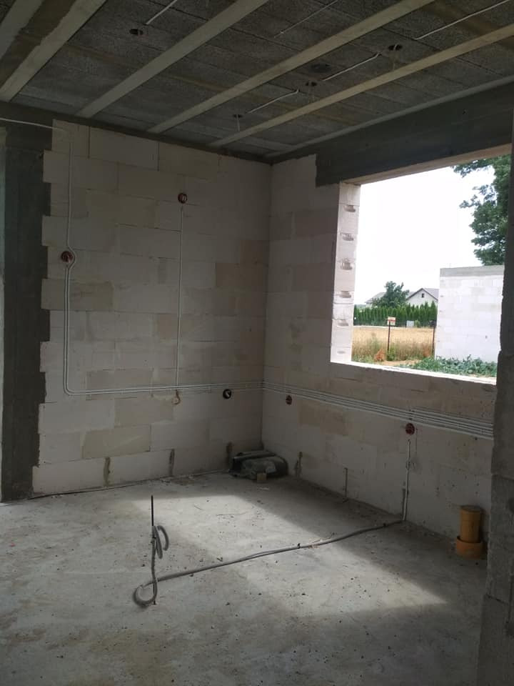 Surowy stan wnętrza z widocznymi przewodami elektrycznymi poprowadzonymi po ścianach z betonu komórkowego, widoczne puszki elektryczne i otwór okienny z widokiem na zewnątrz.