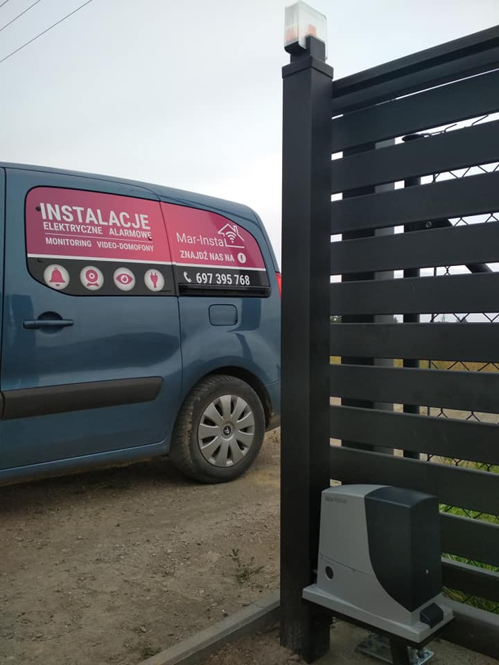 Błękitny samochód dostawczy z reklamą usług elektrycznych i alarmowych firmy Mar-Instal, zaparkowany obok czarnego, nowoczesnego ogrodzenia z zamontowanym napędem bramy.