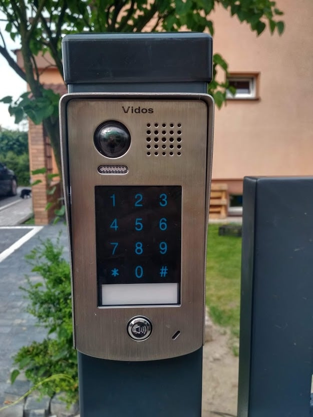 Zewnętrzny panel domofonu Vidos z klawiaturą numeryczną i kamerą, zamontowany na słupku bramy wjazdowej, widok z bliska na tle zieleni i budynku.