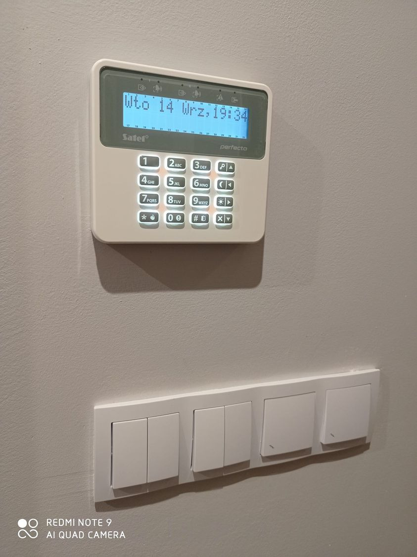 Biały panel sterowania alarmem Satel Perfecto, zamontowany na szarej ścianie, widoczne cztery białe włączniki światła poniżej.