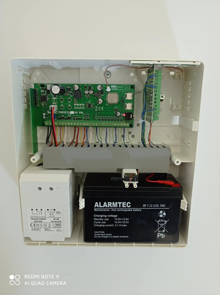 Otwarta obudowa centrali alarmowej z widoczną płytą główną, transformatorem i akumulatorem podtrzymującym marki Alarmtec, z podłączonymi przewodami.