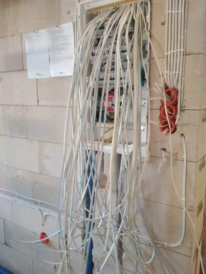 Rozdzielnica elektryczna w trakcie montażu, z licznymi przewodami wychodzącymi z puszki, ściana z bloczków betonowych w surowym stanie, widoczne peszle i kable rozprowadzone po ścianie.