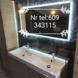 Nowoczesna łazienka z podwójną umywalką, podświetlanym lustrem z numerem telefonu i marmurowymi płytkami na ścianie.