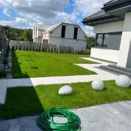 Nowoczesny ogród z równo przystrzyżonym trawnikiem, ścieżką z jasnych płyt, szarymi kulami dekoracyjnymi i zwiniętym zielonym wężem ogrodowym na kostce brukowej, w tle dom w trakcie budowy.