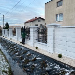 Green Garden Usługi Brukarskie Monika Wagner - Nowoczesne ogrodzenie z białych bloczków betonowych z elementami kutymi, zabezpieczenie skarpy folią i kamieniami, małe tuje wzdłuż ogrodzenia, pracownik w zielonej kurtce przy bramie.
