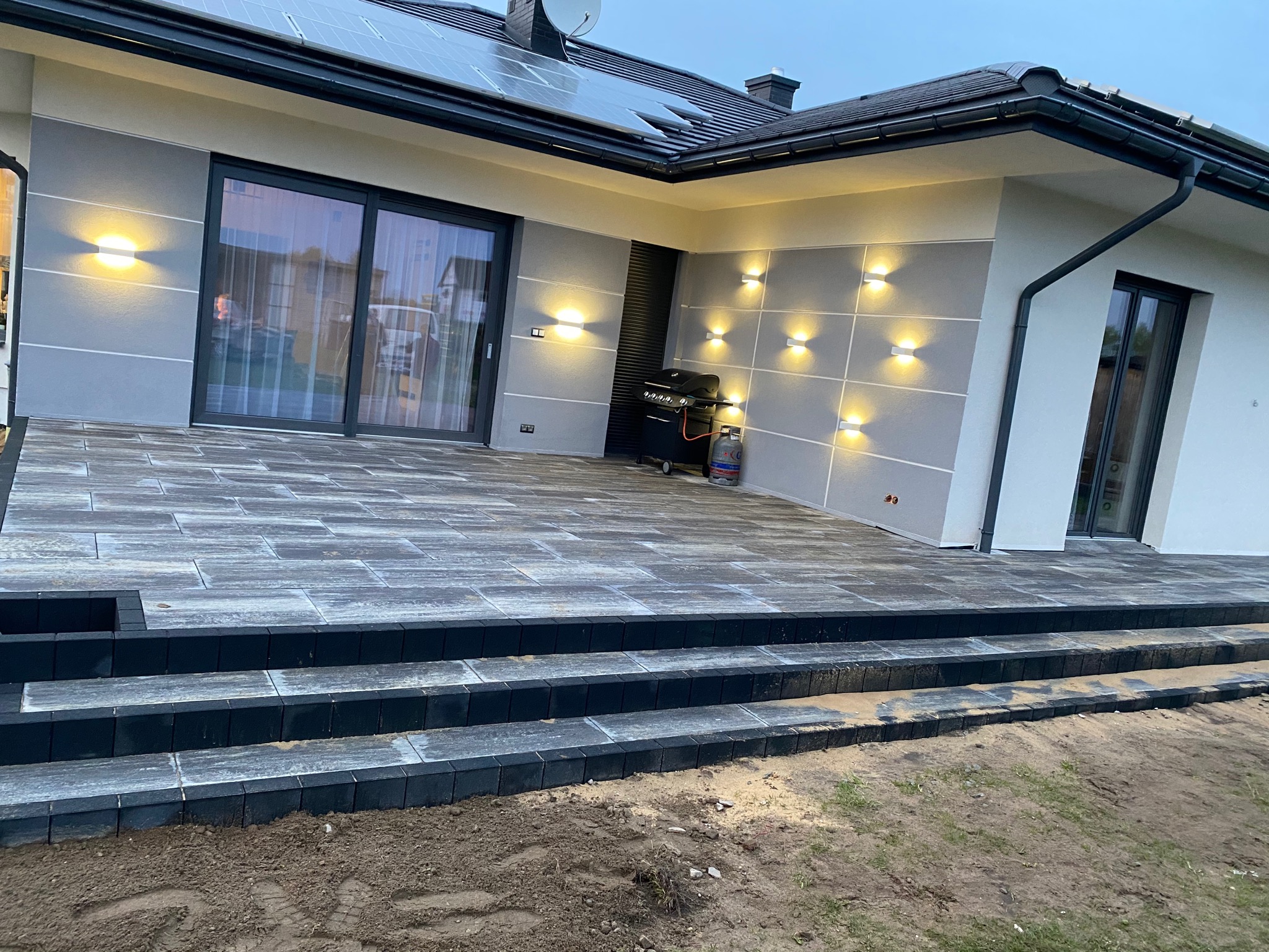 Nowoczesny taras z kostki brukowej w odcieniach szarości, z trzema stopniami i oświetleniem elewacyjnym, grill gazowy na tle ściany.