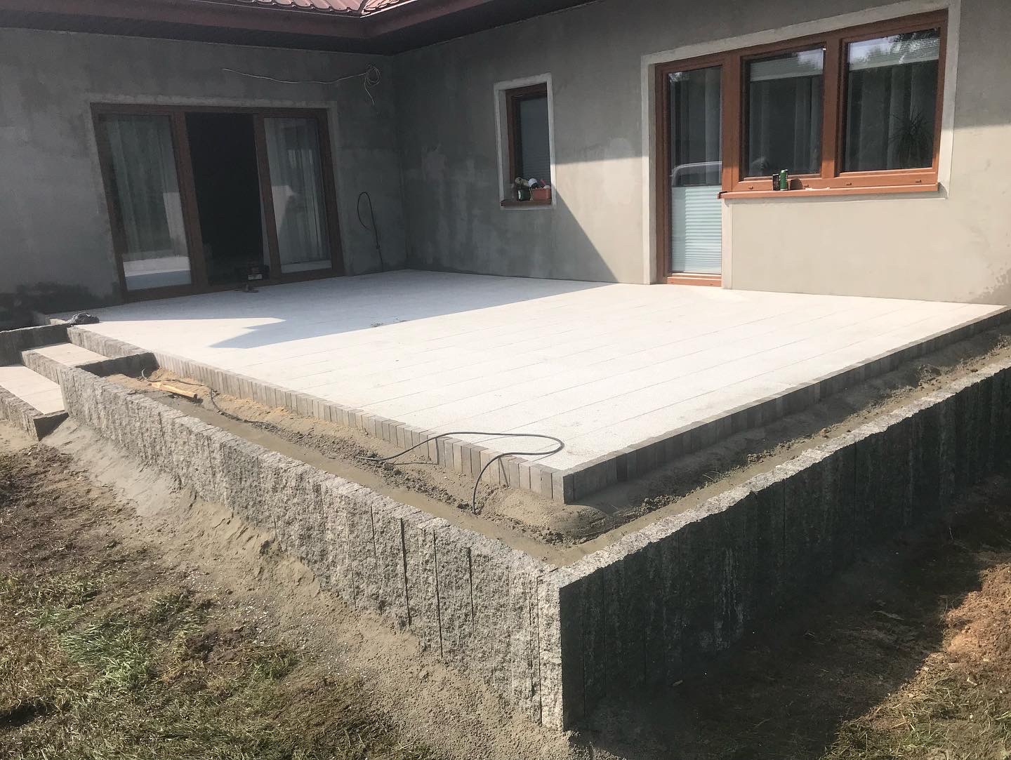 Nowo wyłożony, jasny taras z kostki na podwyższeniu, otoczony obrzeżem z pionowo ułożonych, szarych elementów kamiennych. Widoczne schody z kostki prowadzące na taras, kabel elektryczny na obrzeżu...