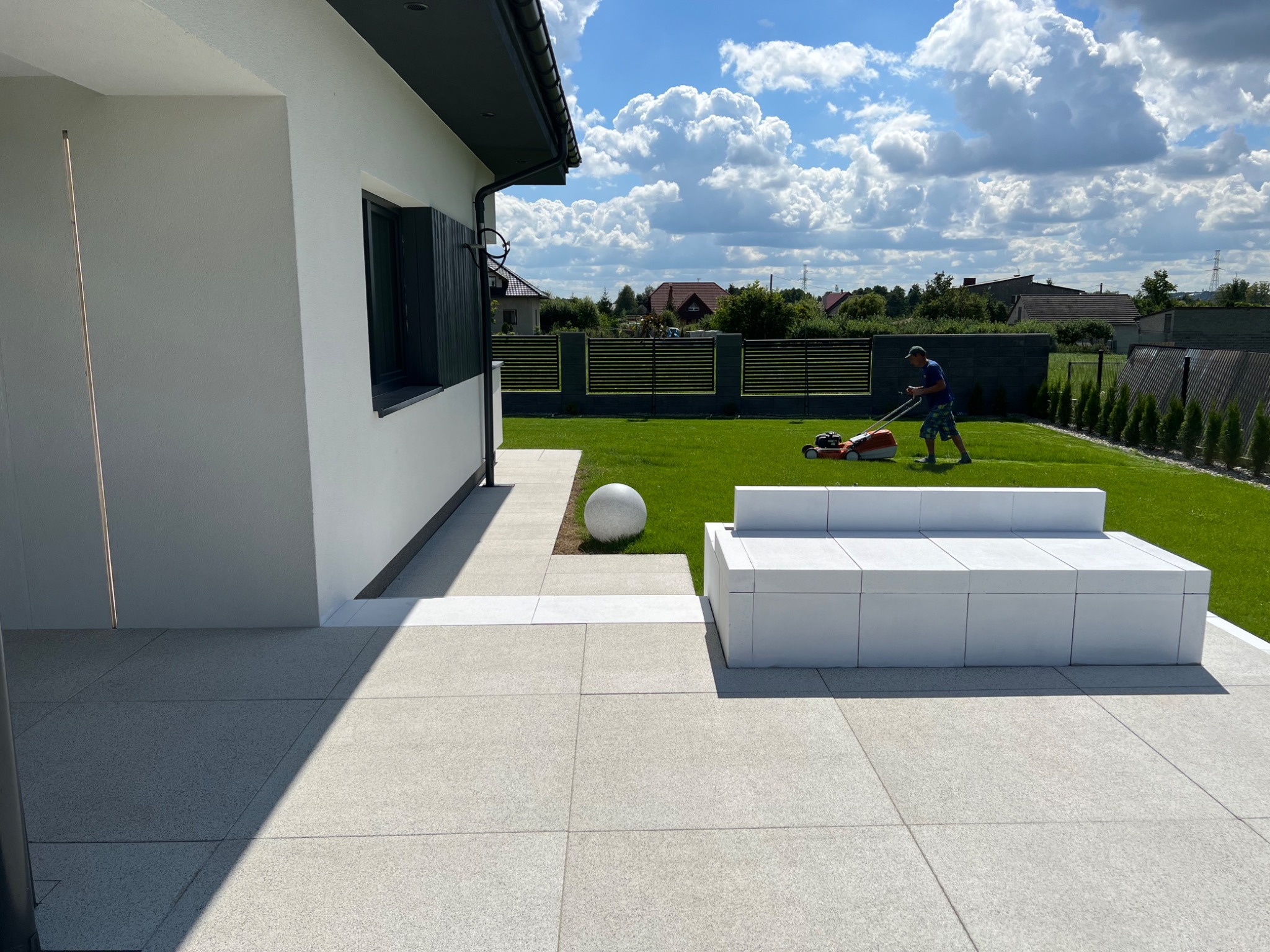 Nowoczesny ogród z jasną kostką, białym siedziskiem i trawnikiem, na którym mężczyzna kosi trawę kosiarką spalinową, w tle widoczny dom i ogrodzenie.