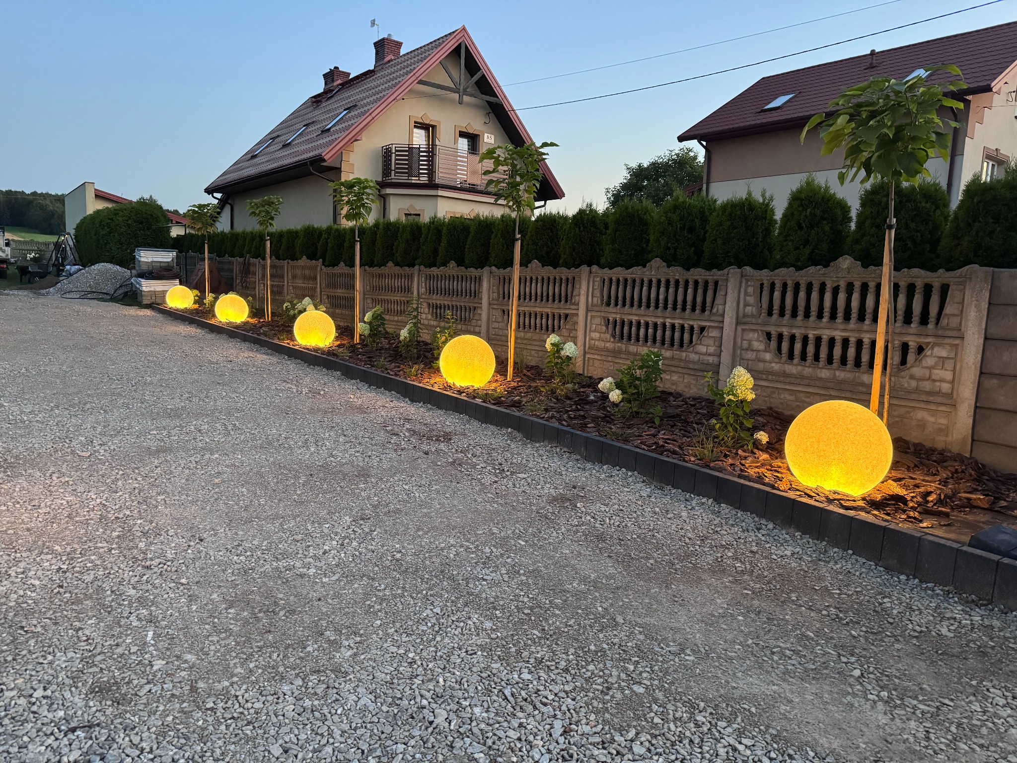 Oświetlona żwirowa droga dojazdowa z kulistymi lampami wzdłuż betonowego ogrodzenia i świeżo posadzonej zieleni.