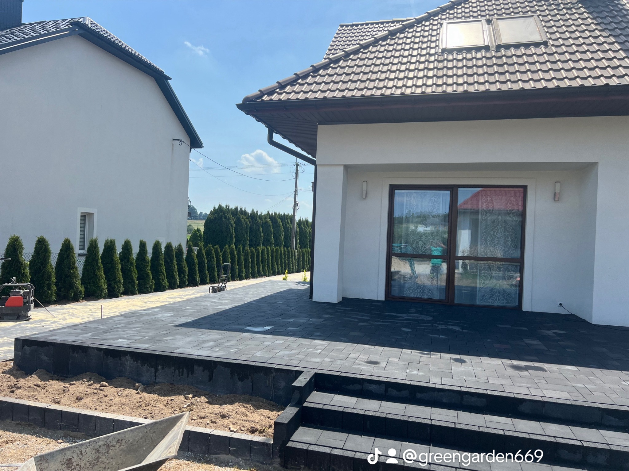 Nowo ułożony, ciemnoszary taras z kostki brukowej przed nowoczesnym domem z brązowym dachem, widoczne schody z kostki, w tle rząd tuj i budynek sąsiada.