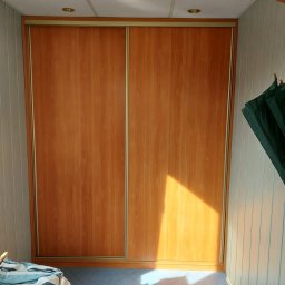 Firma usługowa MARFI - Wnętrze pokoju z zabudowaną szafą przesuwną o drewnopodobnych frontach, oświetlonymi promieniami słońca, z widocznym fragmentem sufitu kasetonowego i niebieskiej podłogi z płytek, oraz fragmentem...