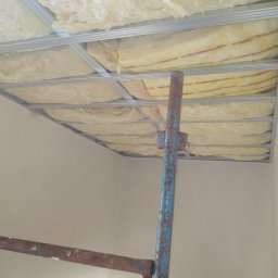 Firma usługowa MARFI - Montaż sufitu podwieszanego z widoczną konstrukcją metalową i ociepleniem z wełny mineralnej, wsparty na niebiesko-rdzawym rusztowaniu.