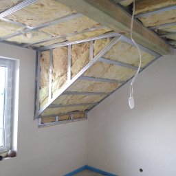 Firma usługowa MARFI - Skośny sufit podwieszany w trakcie montażu, widoczne metalowe profile i wełna mineralna, okno z parapetem w tle, zabezpieczona podłoga.