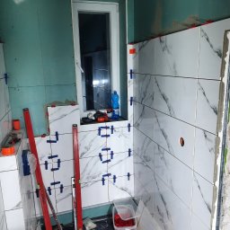 Firma usługowa MARFI - Wnętrze łazienki w trakcie układania białych płytek imitujących marmur, widoczne poziomice, plastikowe krzyżyki dystansowe, niezabudowana ściana z płyty gipsowo-kartonowej oraz okno.