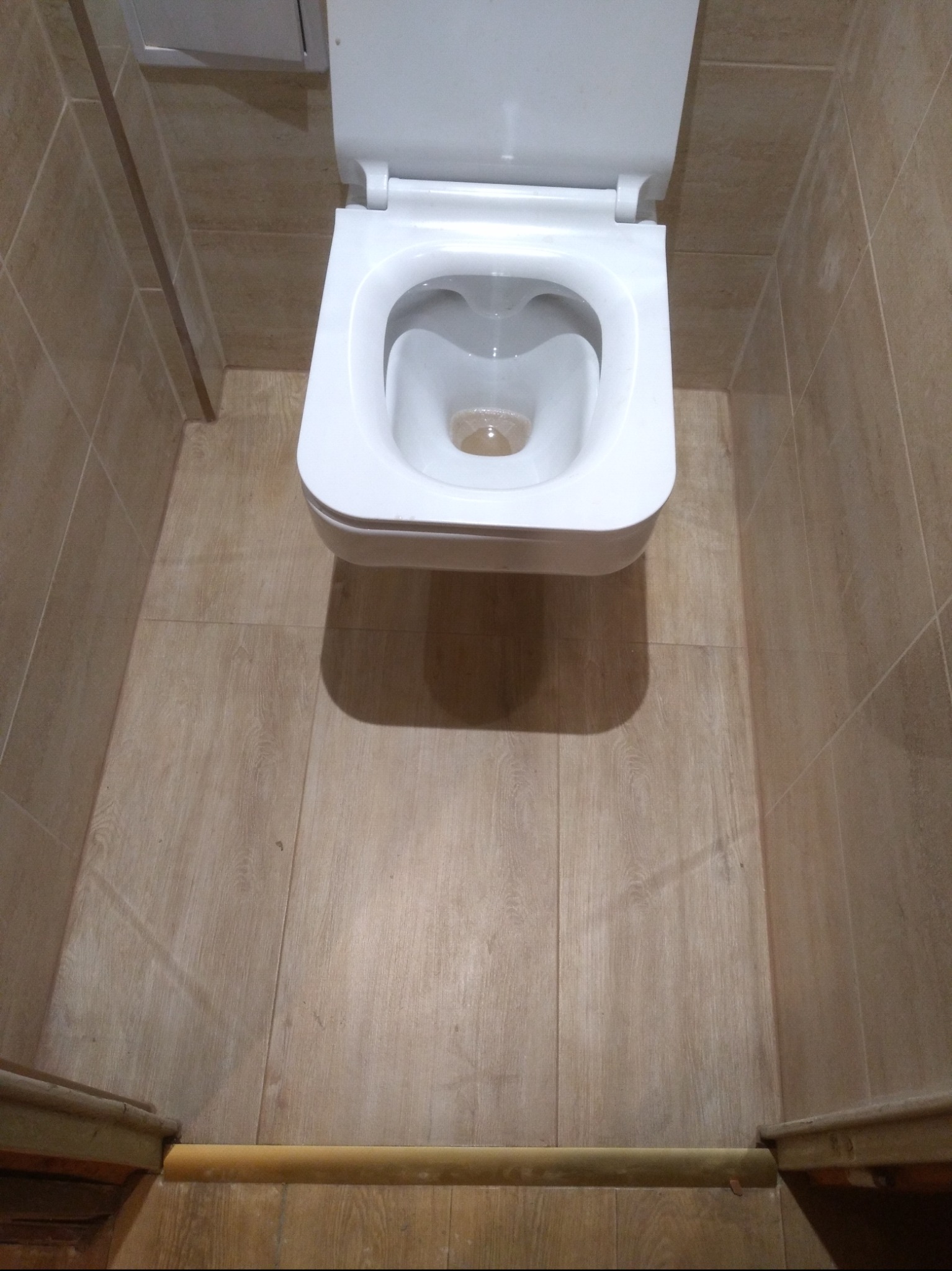 Ceramiczna toaleta wisząca z otwartą klapą, zamontowana w wykończonej łazience z beżowymi płytkami na ścianach i podłodze imitującej drewno, widok z góry.