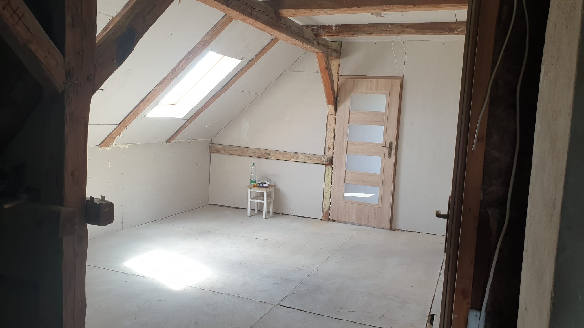 Adaptacja poddasza: surowe ściany z płyt karton-gips, drewniane belki stropowe, okno dachowe i częściowo położona podłoga. Widoczny biały taboret z butelką wody.