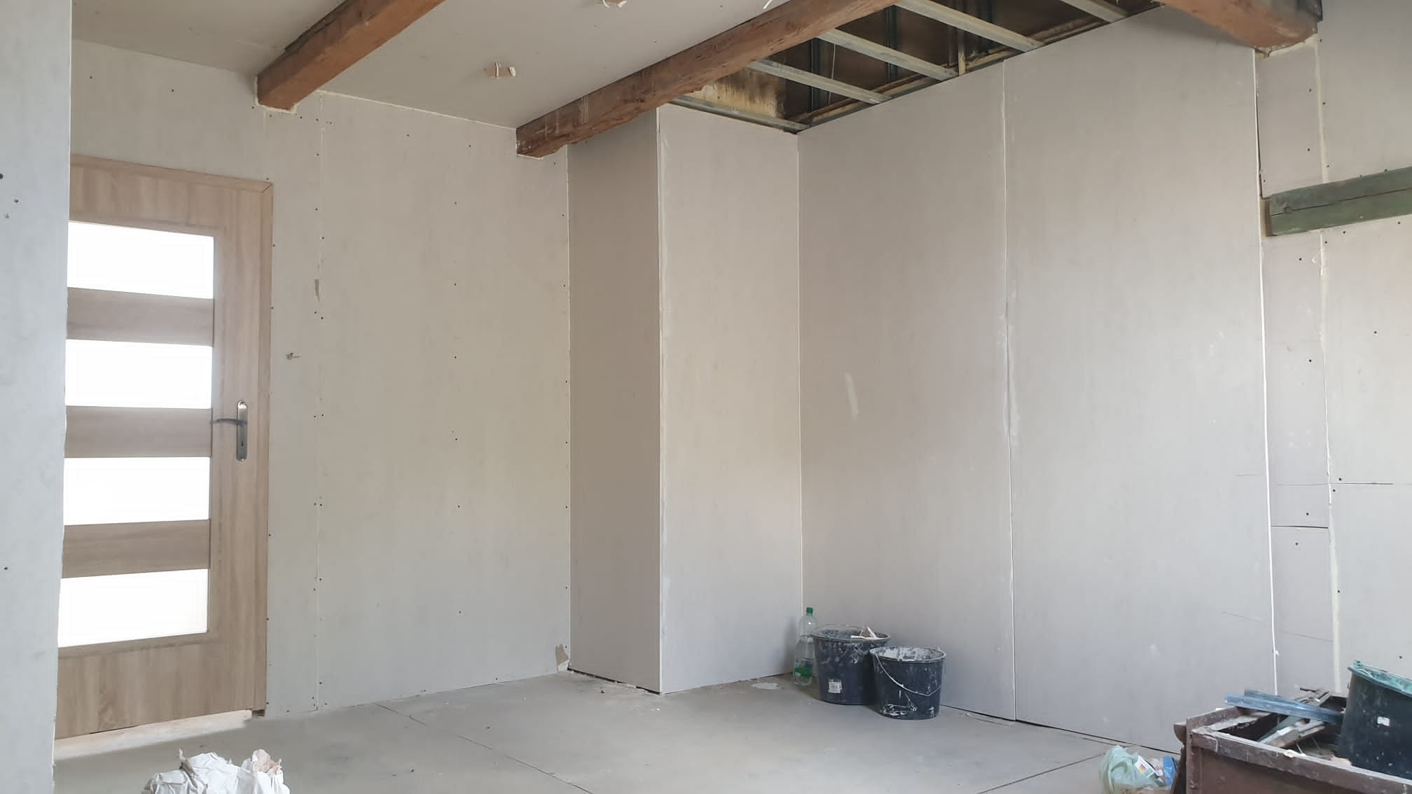 Wnętrze w trakcie prac wykończeniowych: ściany pokryte płytami gipsowo-kartonowymi, widoczne drewniane belki stropowe, drzwi z przeszkleniami, dwa wiadra i butelka wody w rogu pomieszczenia, surowy...