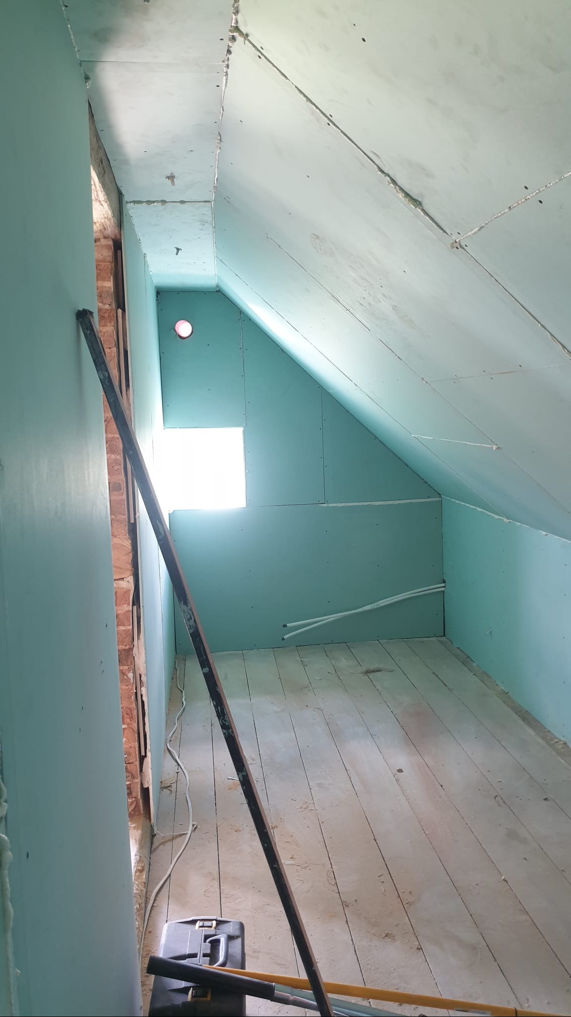 Poddasze w trakcie wykańczania płytami gipsowo-kartonowymi, widoczne narzędzia i skrzynka narzędziowa, częściowo odsłonięta cegła, surowe drewniane deski podłogowe.