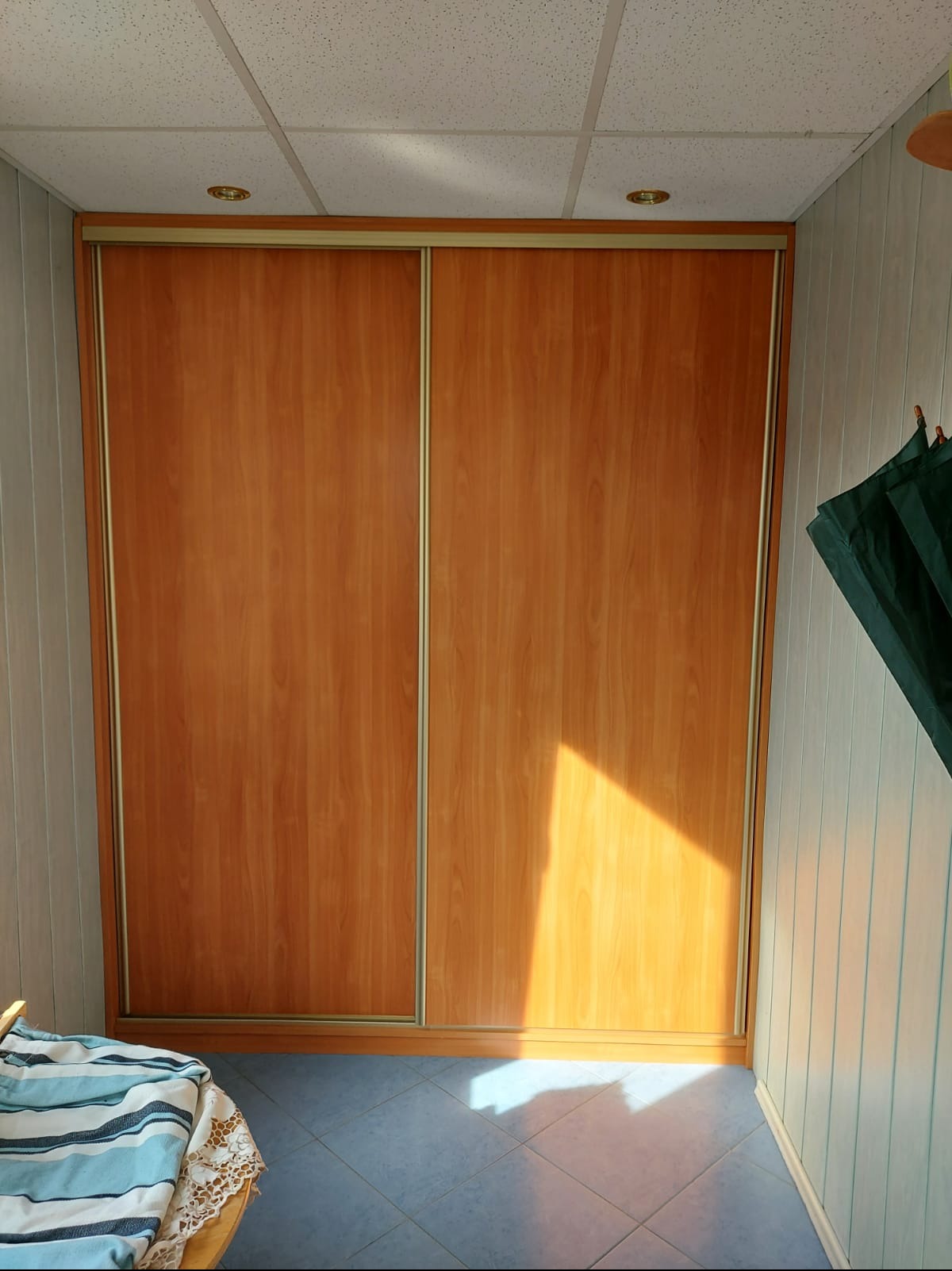 Wnętrze pokoju z zabudowaną szafą przesuwną o drewnopodobnych frontach, oświetlonymi promieniami słońca, z widocznym fragmentem sufitu kasetonowego i niebieskiej podłogi z płytek, oraz fragmentem...