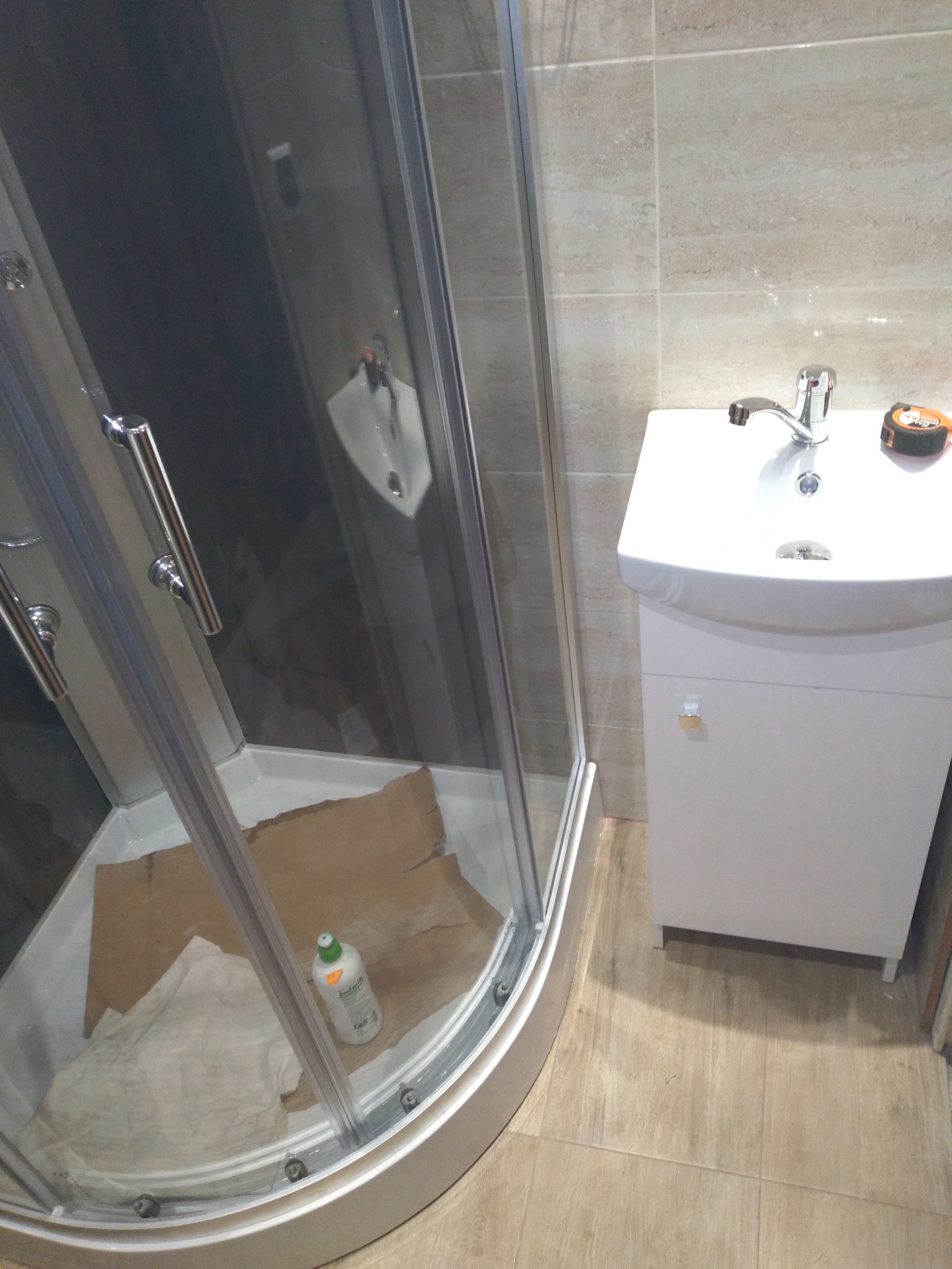 Narożna kabina prysznicowa z zabezpieczeniem podłogi kartonem i butelką płynu do czyszczenia w środku, obok umywalka z szafką i baterią, oraz miarka zwijana na blacie.