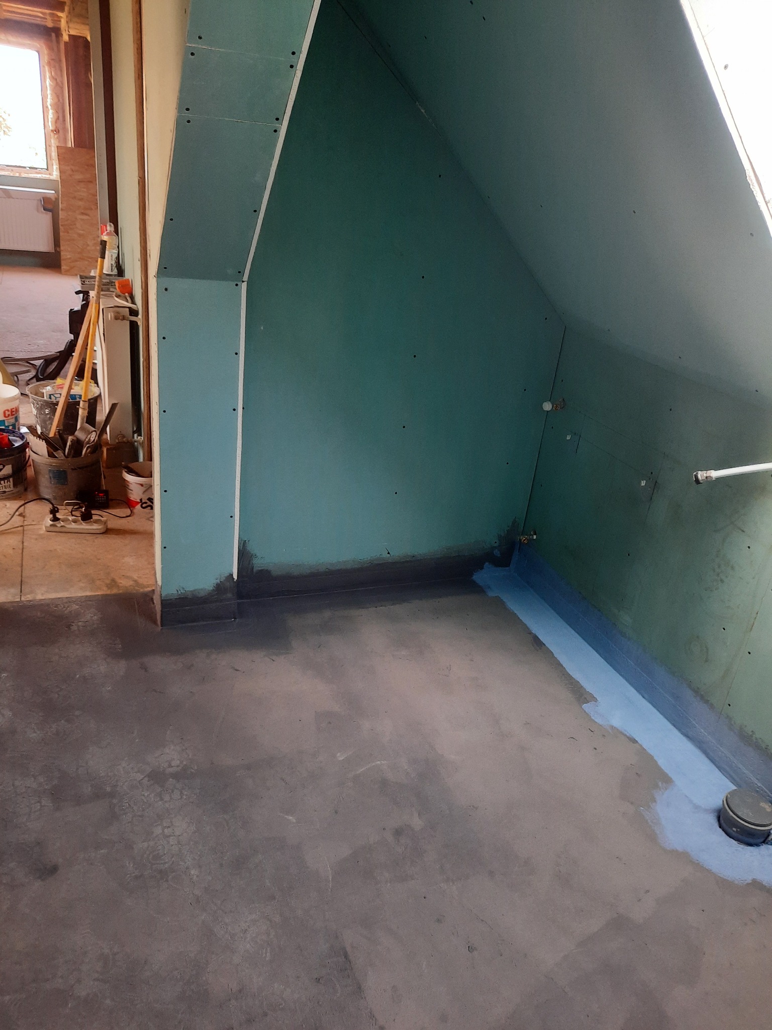 Adaptacja poddasza: ściany wykończone płytami kartonowo-gipsowymi, zabezpieczone hydroizolacją w narożnikach i przy podłodze, widoczny odpływ kanalizacyjny w posadzce.