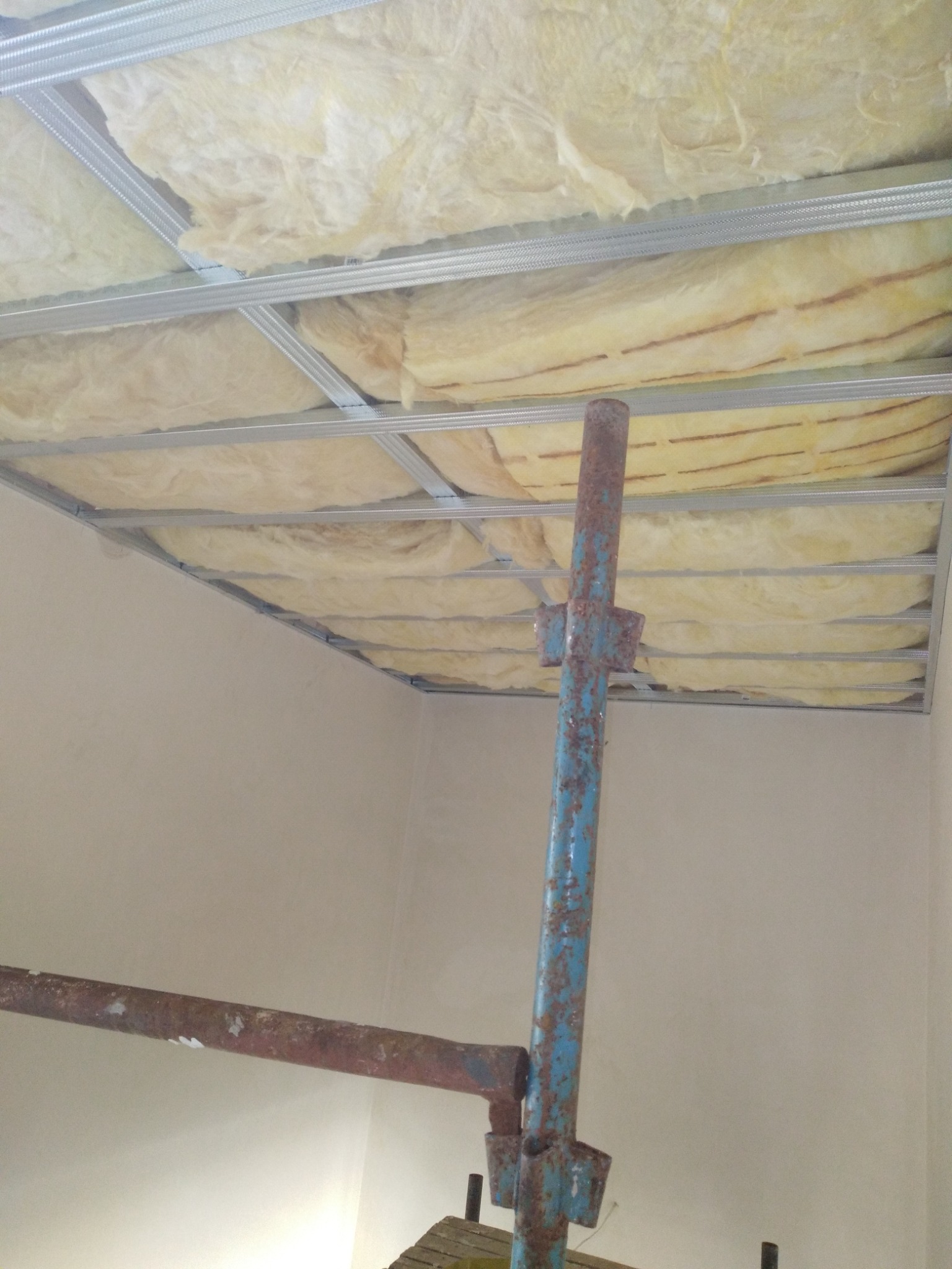 Montaż sufitu podwieszanego z widoczną konstrukcją metalową i ociepleniem z wełny mineralnej, wsparty na niebiesko-rdzawym rusztowaniu.