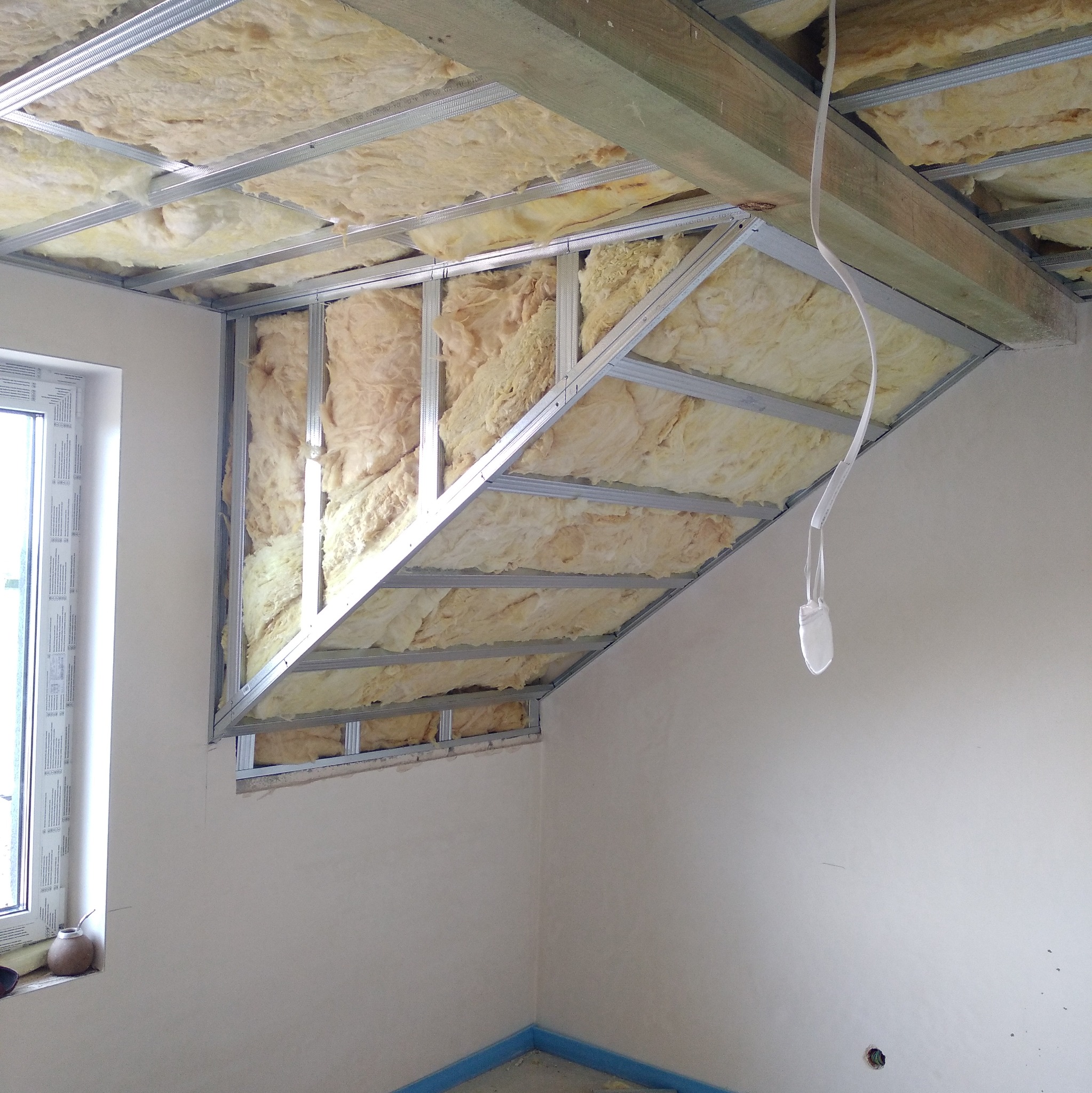 Skośny sufit podwieszany w trakcie montażu, widoczne metalowe profile i wełna mineralna, okno z parapetem w tle, zabezpieczona podłoga.