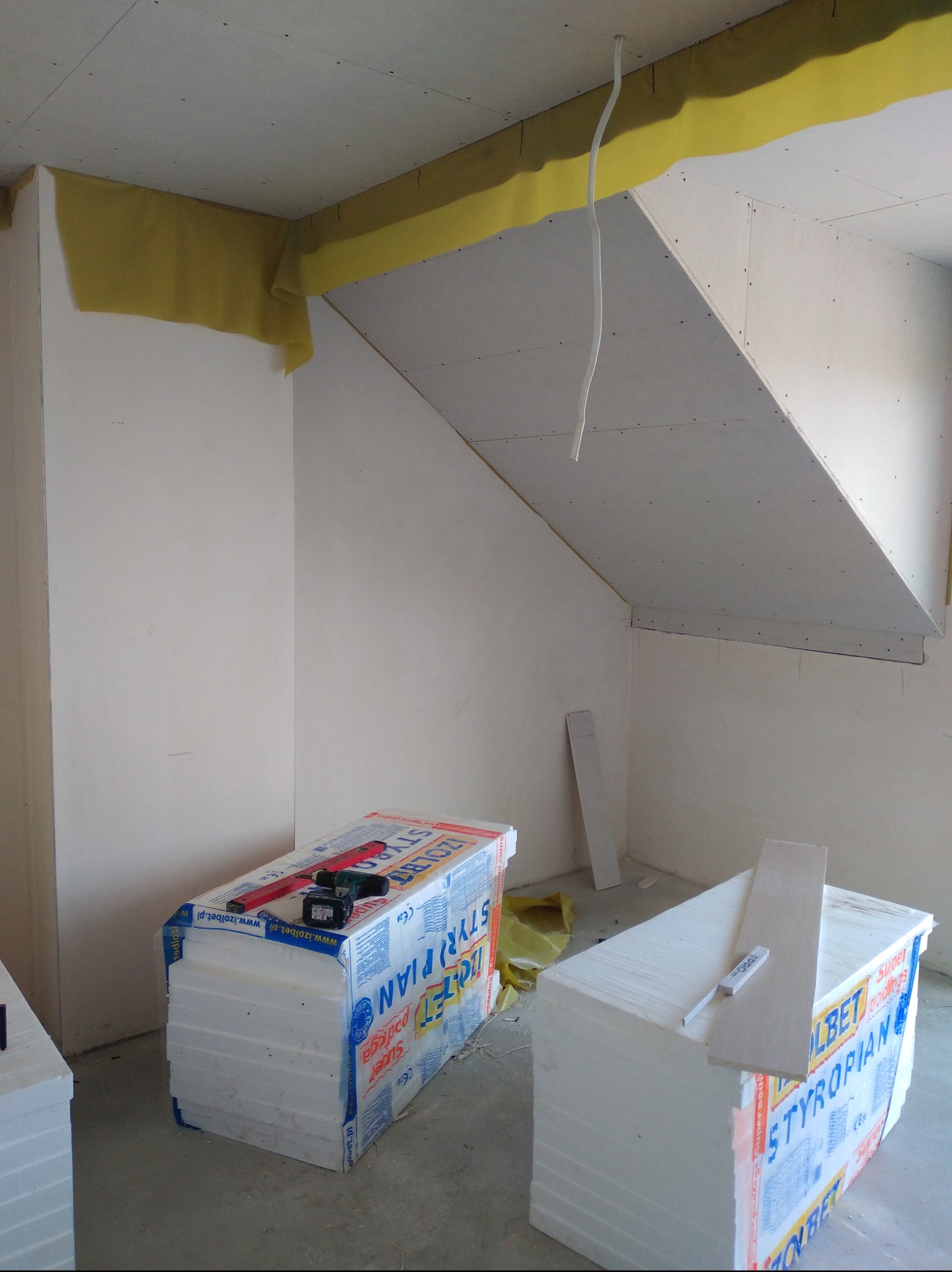 Poddasze w trakcie izolacji: widoczne płyty kartonowo-gipsowe na skosach, żółta wełna mineralna przy suficie, paczki styropianu na podłodze z wkrętarką i poziomicą na jednej z nich.